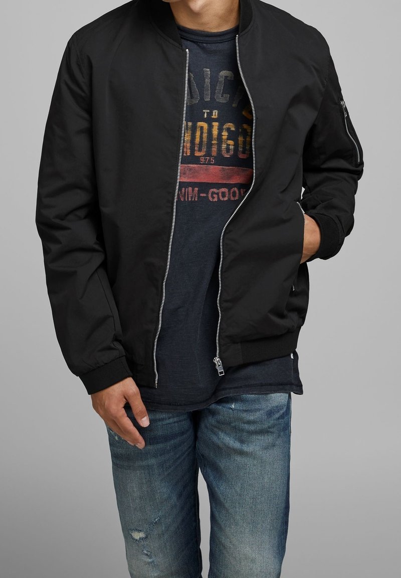 Svart bomberjacka med ribbade muddar och krage, med en silverfärgad dragkedja och sidofickor. Bärs över en mörk t-shirt med grafik och jeans.