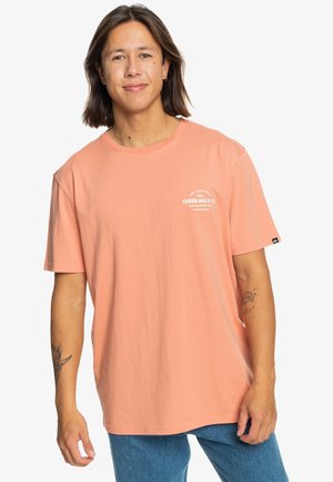Peach-colored cotton T-shirt with short sleeves, featuring a logo design on the chest and a relaxed fit. Worn with blue denim jeans.