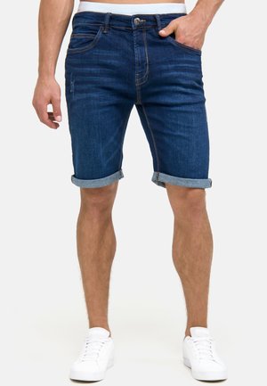 Homme portant un short en jean bleu foncé retroussé et des baskets blanches, debout avec une main dans la poche, sur un fond uni.