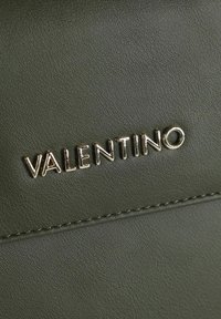 Superficie in pelle verde oliva con finitura testurizzata, caratterizzata da un logo "VALENTINO" in tono oro. Cuciture visibili lungo i bordi.
