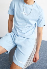 Personne portant un t-shirt et un short light blue de la marque ellesse, assise sur une valise argentée texturée sur un sol recouvert de moquette bleue.