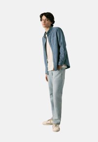 Ljusblå skjorta över en vit T-shirt, kombinerad med ljusa denimjeans och beige sneakers. Modellen står avslappnat med händerna i fickorna.