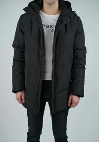 Veste en duvet noire avec capuche, dotée d'une fermeture éclair sur le devant et de boutons-pression, texture matelassée et coupe décontractée, portée sur une chemise blanche.