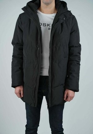 Veste en duvet noire avec capuche, dotée d'une fermeture éclair sur le devant et de boutons-pression, texture matelassée et coupe décontractée, portée sur une chemise blanche.