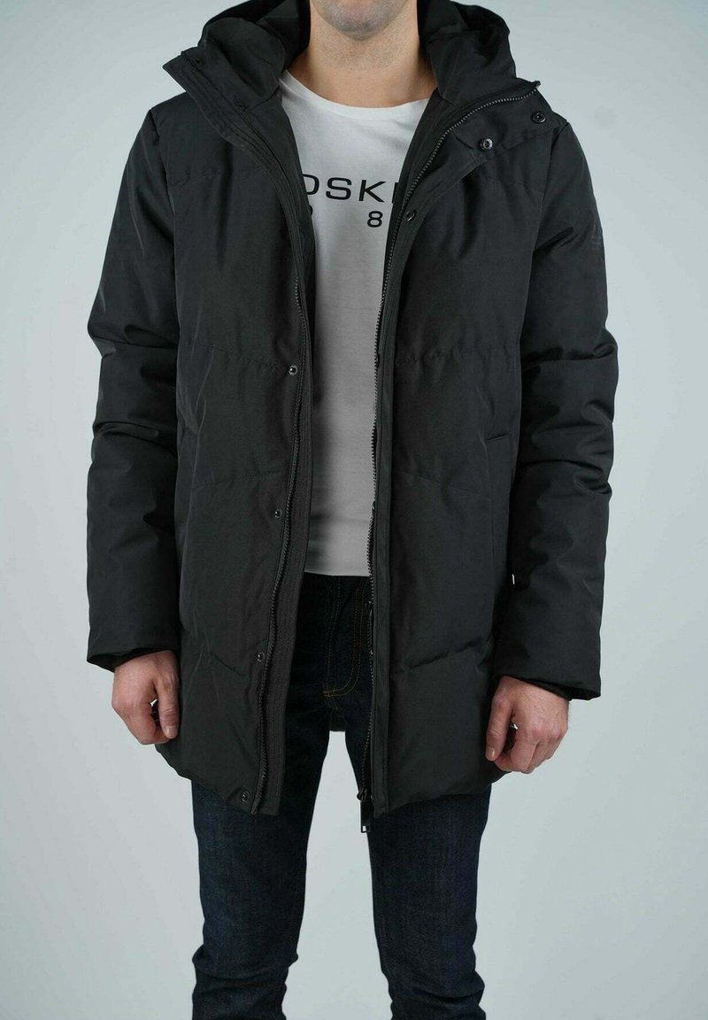 Veste en duvet noire avec capuche, dotée d'une fermeture éclair sur le devant et de boutons-pression, texture matelassée et coupe décontractée, portée sur une chemise blanche.