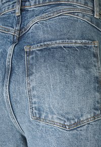 Detailní záběr na zadní kapsu světle modrých džín s viditelným oranžovým prošíváním a texturálními detaily.