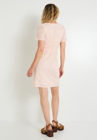 Robe en dentelle de couleur rose doux avec des manches courtes à bords festonnés. Design ajusté, longueur mi-cuisse, assortie à des sandales à plateforme marron.