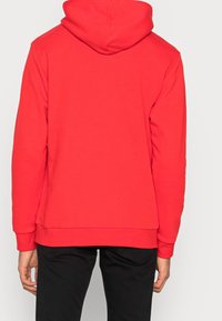 Person som bär en enkel röd huva tröja, sedd från ryggen, kombinerad med svarta byxor mot en enfärgad bakgrund.