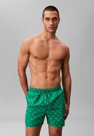 Mężczyzna bez koszulki z krótkimi włosami, ubrany w zielone krótkie spodenki kąpielowe z białym powtarzającym się wzorem logo, stojący na tle jednolitego szarego tła.
