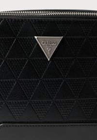 Μαύρο πορτοφόλι από δερματίνη με υφή, με γεωμετρικό μοτίβο τριγώνων και ασημένια πλακέτα με λογότυπο. Διαθέτει κλείσιμο με φερμουάρ για ασφάλεια.