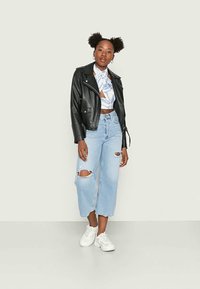 Casaco de motoqueiro de couro preto sobre um top cropped branco e azul com padrão, combinado com jeans largos de ganga azul claro rasgados e sapatilhas brancas.