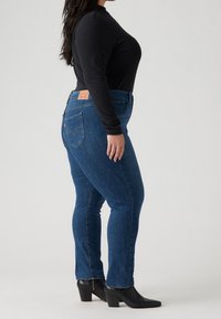 Jeans oscuros de corte ajustado, fabricados en denim, con una cintura de altura media, bolsillos traseros y una etiqueta en la parte posterior. Llevados con una blusa negra de manga larga.