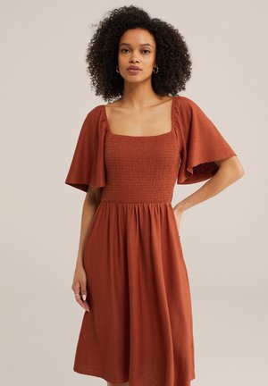 Kohaga lokkis juustega naine, kes kannab ruudukujulise kaeluse ja lendlevate varrukatega roostekarva tekstuurse kleidi, seisab ühe käega puusal.