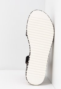 Sandale avec semelle blanche rainurée et lanières noires, positionnée verticalement contre un mur d'angle blanc.