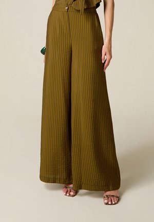 Vrouw draagt olijfgroene wijde gestreepte broek gecombineerd met sandalen met open teen, staande tegen een lichtbeige achtergrond.