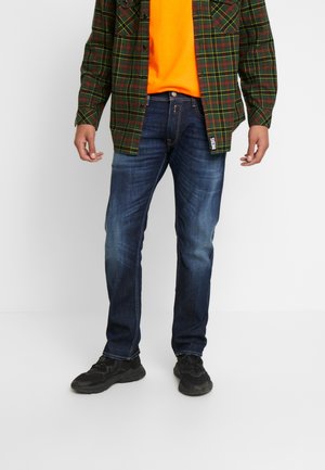 Mies pukeutunut tummansinisiin farkkuihin, mustiin lenkkareihin, kirkkaan oranssiin paitaan ja avoimeen vihreään ruudulliseen nappipaitaan, seisoo valkoisella taustalla.