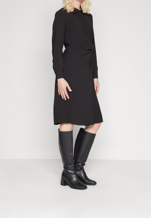 Kobieta w czarnej sukience midi z długim rękawem i zapinanym na guziki kołnierzykiem, w połączeniu z czarnymi skórzanymi kozakami do kolan, stojąca na jednolitym tle.