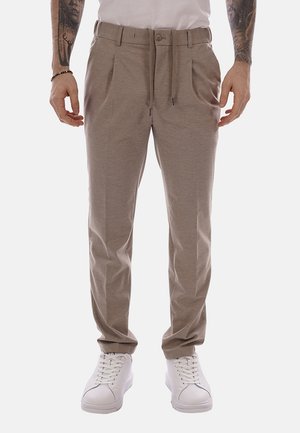 Uomo con pantaloni casual beige con coulisse e bottone, abbinati a scarpe da ginnastica bianche e con le braccia tatuate visibili.