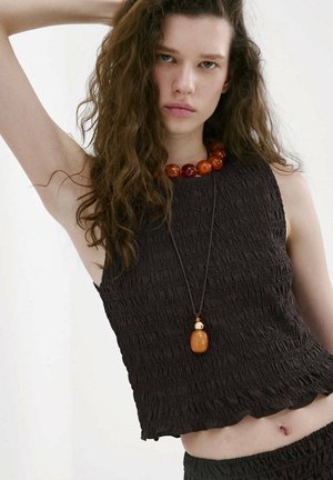 Jeune femme aux longs cheveux bouclés portant un haut noir sans manches texturé, un collier de perles ambre et un pendentif, posant avec un bras levé.