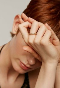 Anillo de oro con detalles delicados, llevado en un dedo delgado, sobre un fondo neutro. El tono de piel es claro y la postura de la mano es relajada.