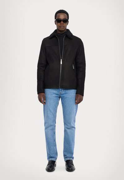 Schwarze Kunstwildlederjacke mit einem Schaffellkragen, Reißverschluss vorne und Seitentaschen, kombiniert mit hellblauen Jeans und schwarzen Schuhen.