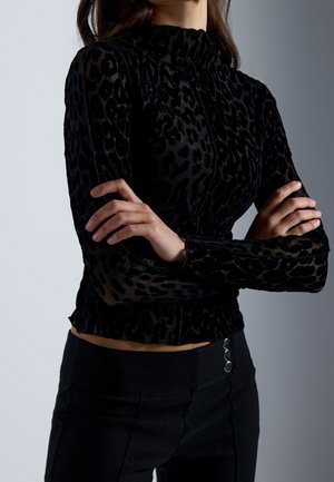 Femme, les bras croisés, porte un haut noir à manches longues avec imprimé léopard et un pantalon noir taille haute à trois boutons.