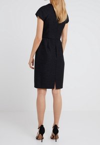 Czarna sukienka z teksturowanym wzorem, krótkimi rękawkami typu cap, dopasowaną talią, długością do kolan oraz rozcięciem z tyłu; noszona z czarnymi sandałami na obcasie.