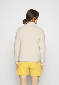 Lys beige fleecejakke med høj krave, blød tekstur og kontrastende kant. Sammen med gule shorts, set fra ryggen.