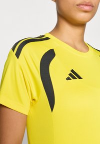 Κίτρινο αθλητικό πουκάμισο με μαύρες λεπτομέρειες και υφή υφάσματος. Διαθέτει στρογγυλή λαιμόκοψη και το λογότυπο  της Adidas στο στήθος, τονίζοντας το σπορ ντιζάιν.