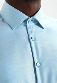 Světle modrá texturovaná košile se strukturovaným límcem, která má tři malé knoflíky ve světlejší modré barvě, a prezentuje tak elegantní a moderní design.