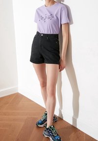 Camiseta de algodón lila con texto bordado en negro, combinada con pantalones cortos de mezclilla negra de tiro alto. El calzado presenta un diseño de zapatillas multicolores.