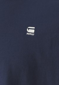 Tmavě modré bavlněné tričko s malým bílým logem "RAW" na levé straně. Materiál má hladkou texturu a klasický design s kulatým výstřihem.