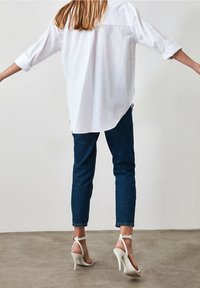 Camisa blanca de manga larga con puños remangados, combinada con jeans de mezclilla azul oscuro. Presenta sandalias de tacón alto blancas, que muestran un diseño minimalista.