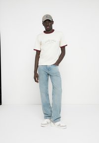 Krém színű rövid ujjú póló bordó díszítéssel, világoskék, széles szárú farmernadrág és bézs színű sportcipő. A modell szürke sapkát visel.