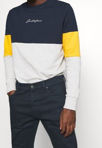 Hombre que lleva una camisa de manga larga en bloques de color con secciones en azul marino, amarillo y gris claro, combinada con pantalones de un azul marino oscuro, con las manos en los bolsillos.