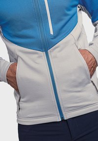 Hellegraue und blaue Zip-Jacke mit strukturiertem Design, ausgestattet mit zwei Seitentaschen und einem Stehkragen. Mit blauen Akzenten am Reißverschluss.