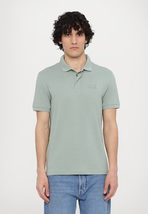 Paita polo made of soft green cotton, featuring a classic collar, short sleeves, and a subtle EA7 logo on the chest. Paired with light blue jeans.