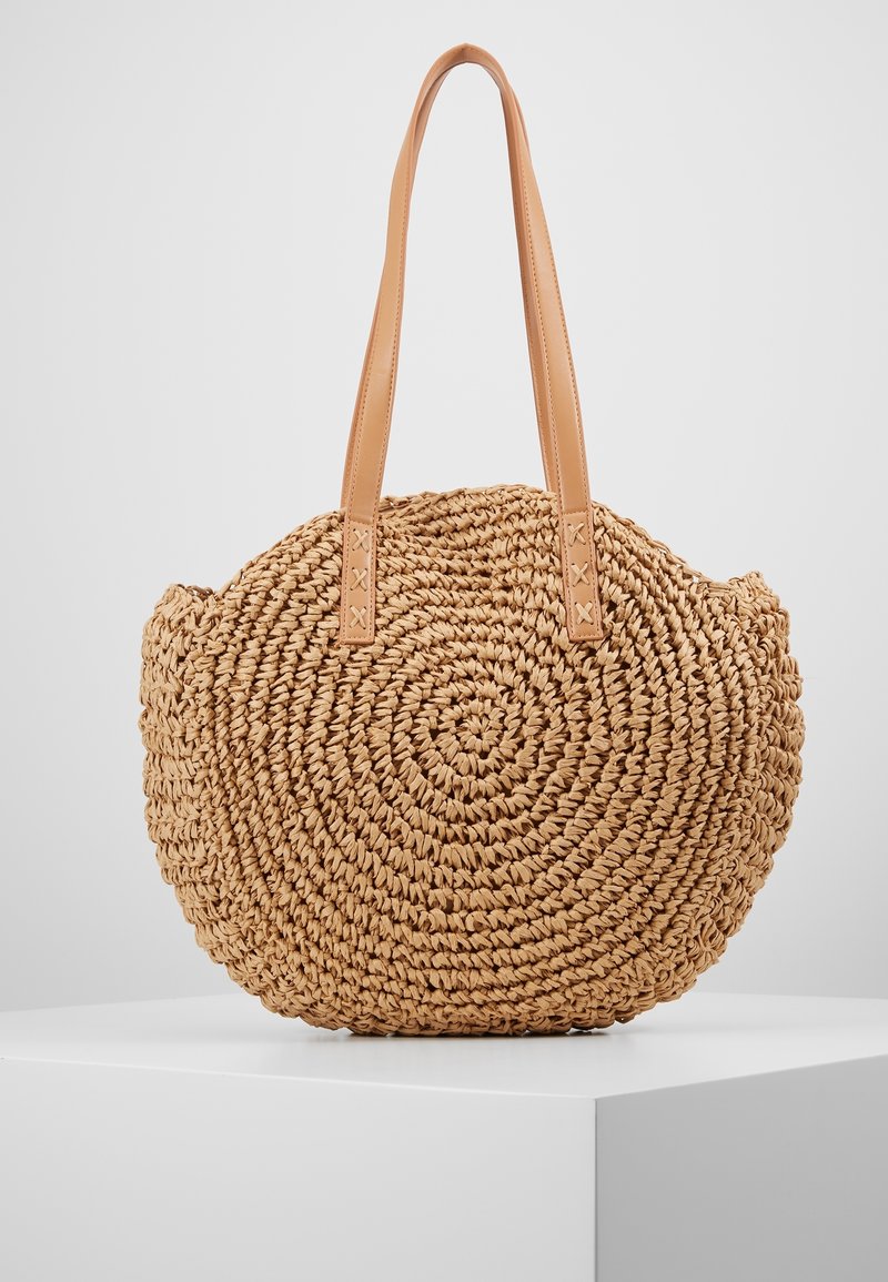 Sac à main rond en paille tissée avec des sangles en cuir marron clair, exposé sur un socle blanc devant un fond gris.