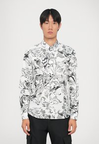 Camicia bianca con motivi astratti neri a spirale, colletto a bottoni, maniche lunghe e dettagli testuali discreti sulla spalla.