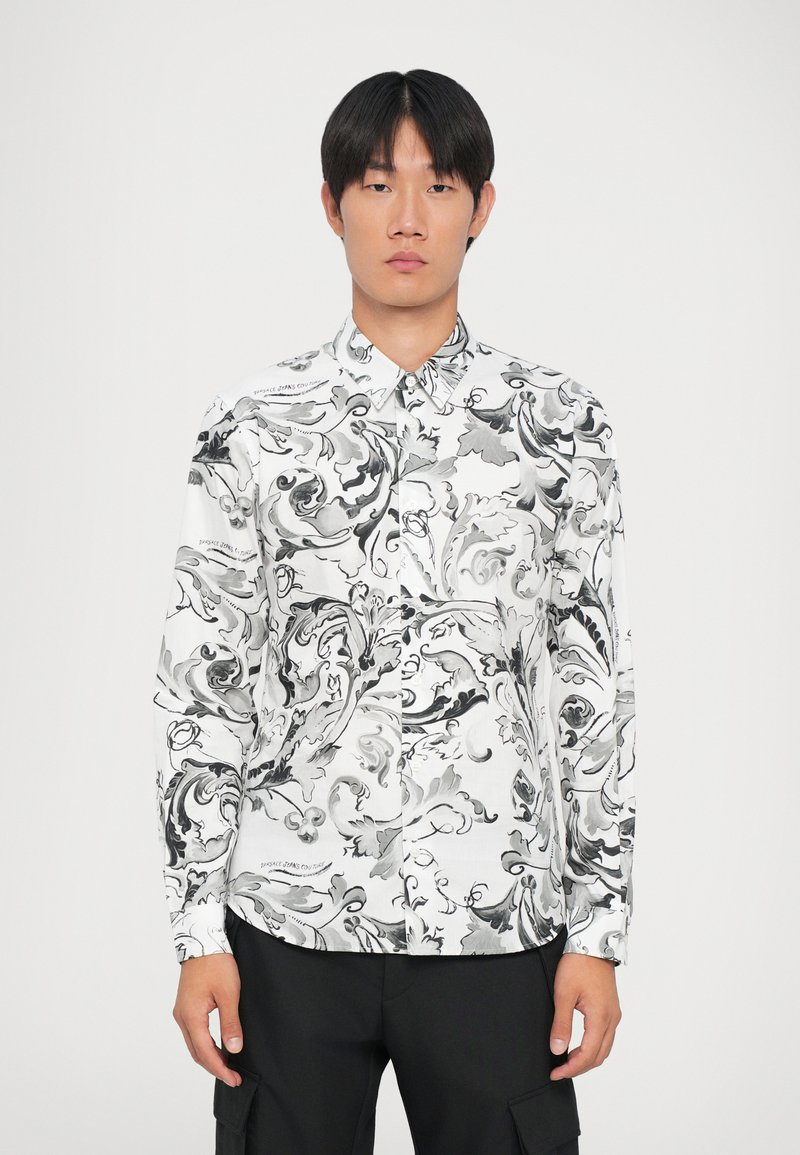 Camicia bianca con motivi astratti neri a spirale, colletto a bottoni, maniche lunghe e dettagli testuali discreti sulla spalla.
