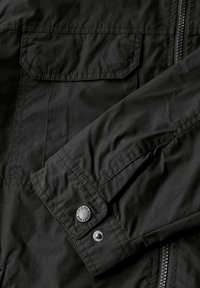 Svart jacka med en slät tygtextur, framfickor med lock, dragkedjestängning och metallknappdetaljer på ärmsluten.