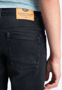 Vaqueros de mezclilla negros con un bolsillo trasero con detalles de costura y una etiqueta de cuero naranja que dice "Denim Driven Since 1989."