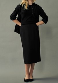 Femme portant une chemise noir à boutons par-dessus un haut noir, une jupe midi noire et des ballerines noires pointues, se tenant avec les mains dans les poches de sa jupe.