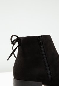 Czarne zamszowe botki za kostkę z bocznym zamkiem błyskawicznym i wiązanym akcentem na pięcie. Posiadają grube czarne podeszwy oraz teksturowany wierzch.