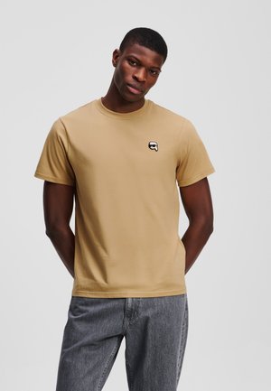 Beige bomulds t-shirt med rund halsudskæring, korte ærmer og et lille sort-hvidt logo på brystet. Båret med grå jeans.