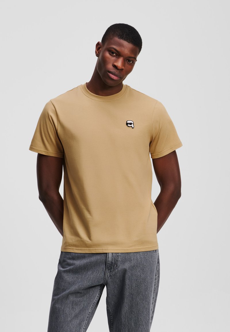 Beige katoenen T-shirt met een ronde hals, korte mouwen en een klein zwart-wit logo op de borst. Draagt een grijze spijkerbroek.