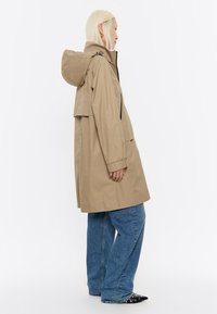 Beige waterdichte jas met capuchon, zijzakken met rits en een relaxte pasvorm, gecombineerd met blauwe wijde jeans en zwarte pumps.
