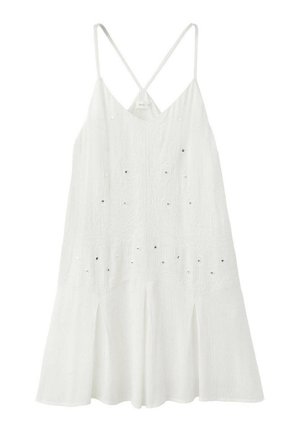 Joven mujer con un vestido blanco sin mangas que sostiene un bolso negro con flecos, lleva sandalias planas marrones con tachuelas, de pie frente a un fondo blanco liso.