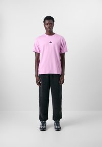 Ljust rosa kortärmad t-shirt med en liten svart logotyp, i kombination med mörka strukturerade fleecebyxor och mönstrade sneakers.