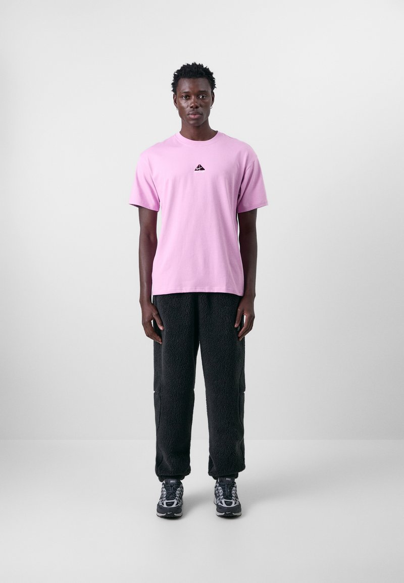 Ljust rosa kortärmad t-shirt med en liten svart logotyp, i kombination med mörka strukturerade fleecebyxor och mönstrade sneakers.